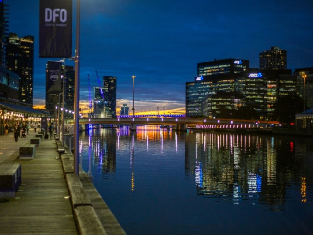 Melbourne South Wharf waterfront at dusk with DFO sign, illuminated bridge and city skyline reflections on Yarra River, Victoria, Australia